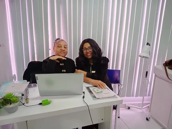 Clínica de Estética no Rio de Janeiro 4 Duas profissionais da saúde da Clínica Sarayu sentadas sorrindo em ambiente acolhedor, ao lado de laptop e prontuários, em uma sala de atendimento moderna.