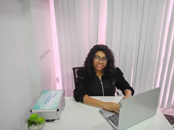 Dra. Suely Gomes da Fonseca, especialista em ozonioterapia, sorrindo enquanto trabalha em seu consultório na Sarayu Estética e Saúde, ao lado de um notebook e maleta de terapias integrativas.